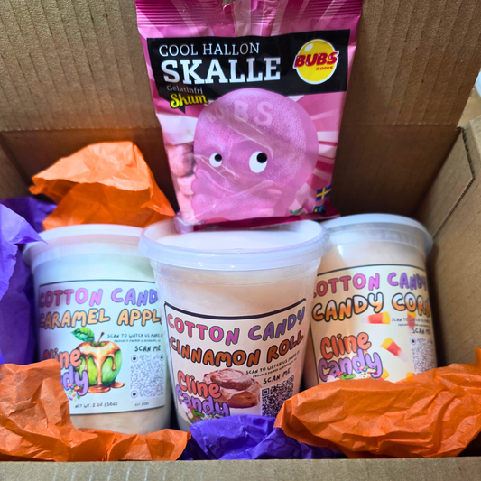 A gift box filled with cotton candy and a bag of Skoomi 'Cool Hallon Skalle' cotton candy. The box contains three white plastic tubs of cotton candy (Cotton Candy Caramel Apple, Cotton Candy Cinnamon Roll, and Cotton Candy Clime Candy) surrounded by crumpled orange and purple tissue paper, with a pink bag of Skoomi cotton candy standing on top.